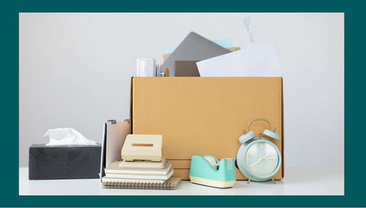 A desk with items being packed - a brown box and next to it a clock, stapler, tissues and notepads