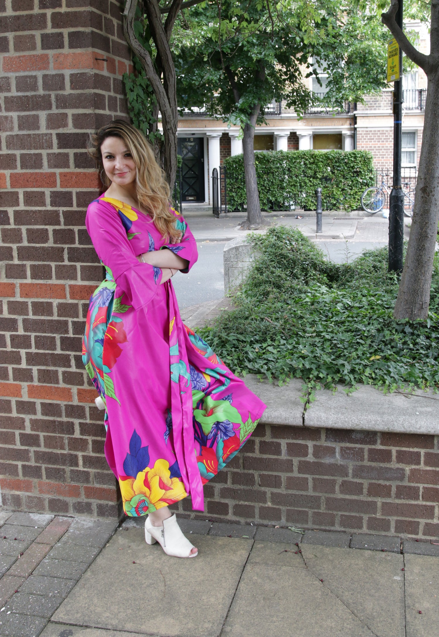 Ioana wearing a strong pink dress with flower prints and leaning against a brick pillar. In the background there are plants and trees