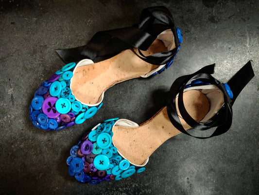 Sandal like flats with open section between vamp (toe area) and back heel panel. The front toe area is covered in buttons with various shades from deep blue, purple and light blue. The back section is covered in dark blue buttons and has a lace silky strap along the ankle to help with support. The inside is leather in a natural tan color