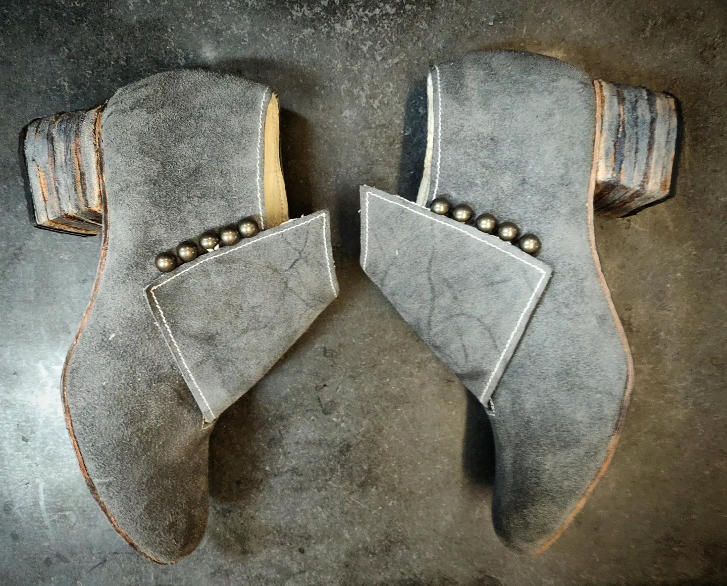 Boots in grey suede leather, a block heel of 7cm also dyed grey. They have a flap over the front area which goes from the inside of the shoe to the outside in a asymmetric manner and is closed with 5 dark grey leather metal buttons with are dome shapdd
