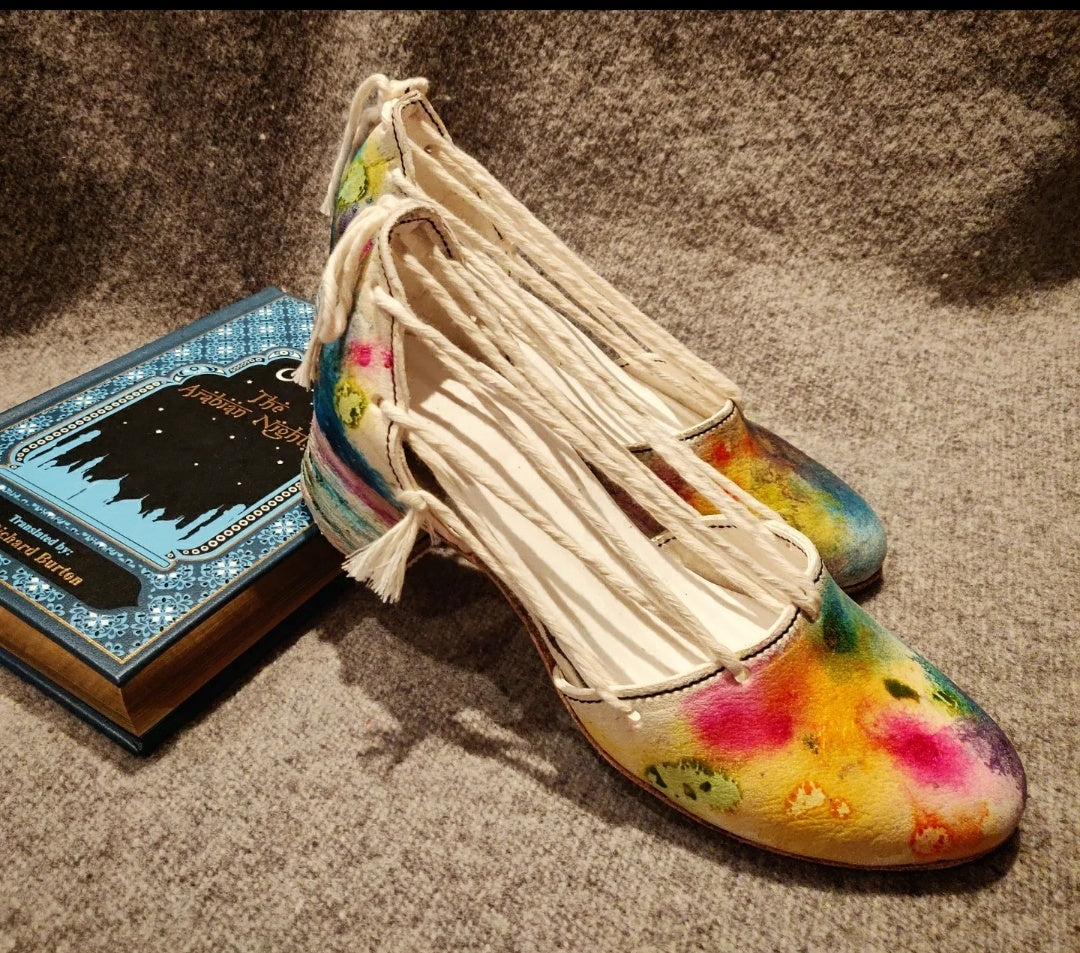 The shoes propped against the book the Arabian Nights. Focus on the front area of the right shoe with vibrant yellow, purples, greens and blues 
